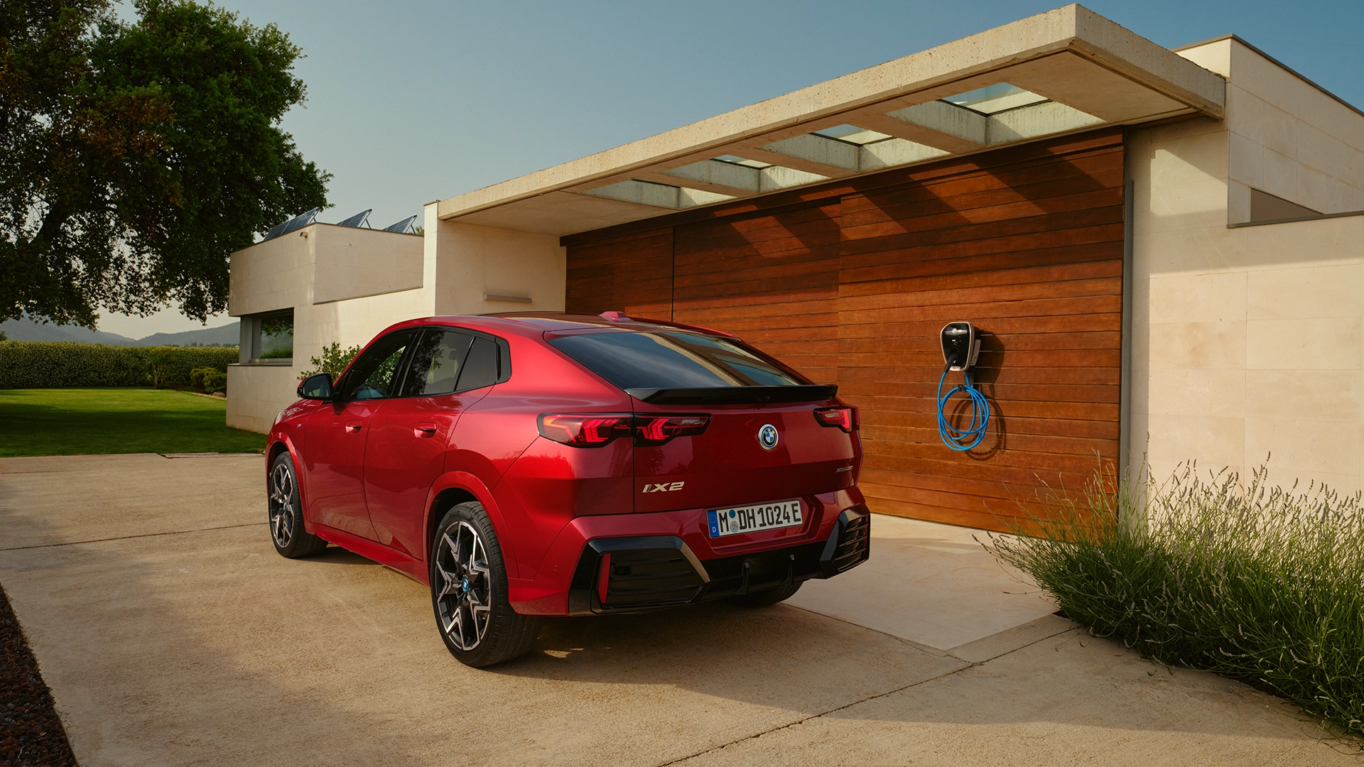 Ein roter BMW iX2 steht in der Einfahrt eines Hauses. Das Haus besitzt eine Ladestation für Elektroautos. Es besitzt ebenso Solarpanelle und einem Garten mit einem großen Baum.