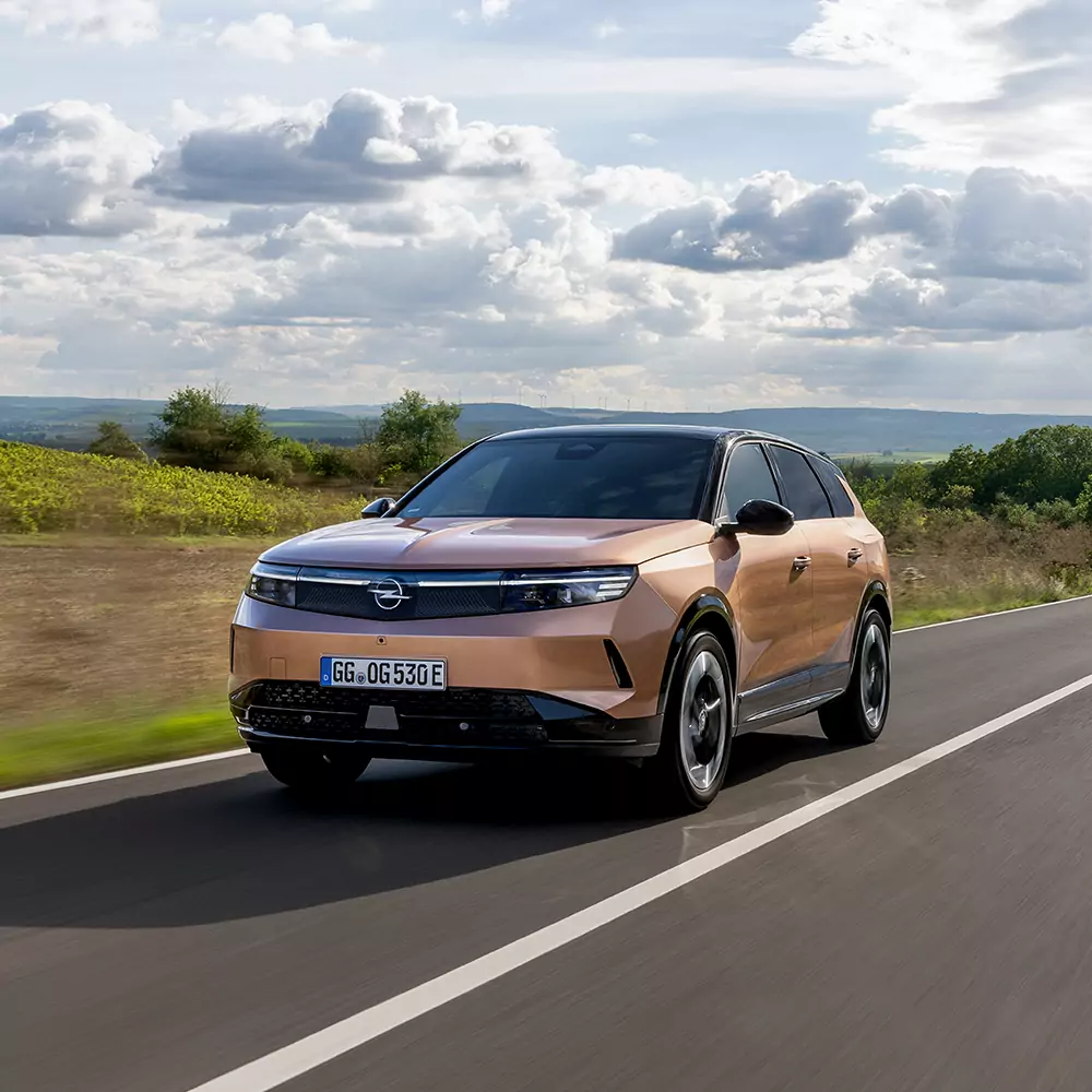 Ein Opel Grandland fährt auf einer Straße mit einer Landschaft im Hintergrund. Im Horizont erkennt man mehrere Windräder. Der Himmel ist bewölkt.
